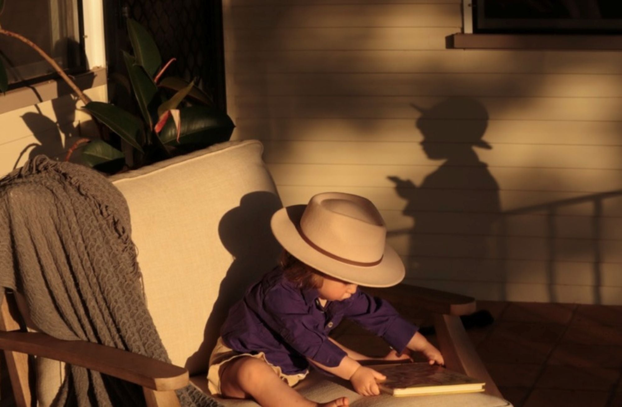An Australian toddler wearing a beige wide-brim fedora hat and a purple shirt sits on a porch chair reading a book, with warm sunlight casting a long shadow on the wall behind them.