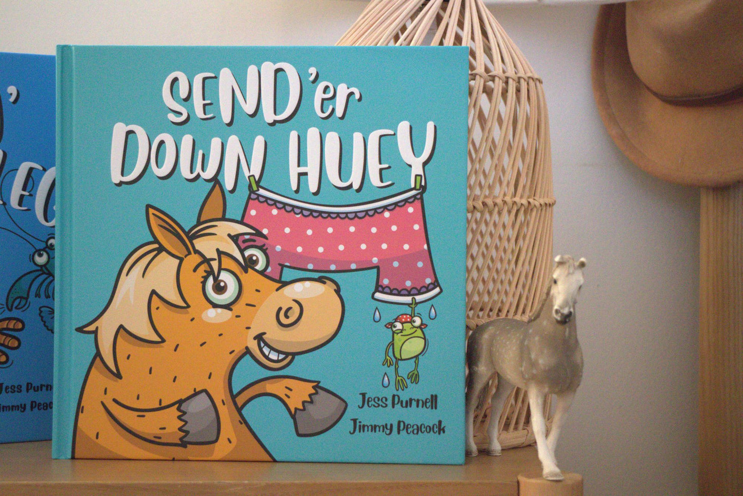 An Australian children's book titled "SEND 'er DOWN HUEY" by Jess Purnell and Jimmy Peacock, featuring a turquoise cover with a cartoon horse and laundry scene, displayed on a shelf next to a small horse figurine and a woven basket.