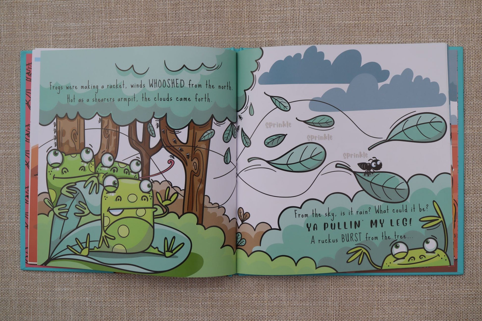An open Australian children's picture book showing a whimsical forest illustration with green frogs, trees, flying leaves, and printed text on both pages, photographed on a beige fabric surface.