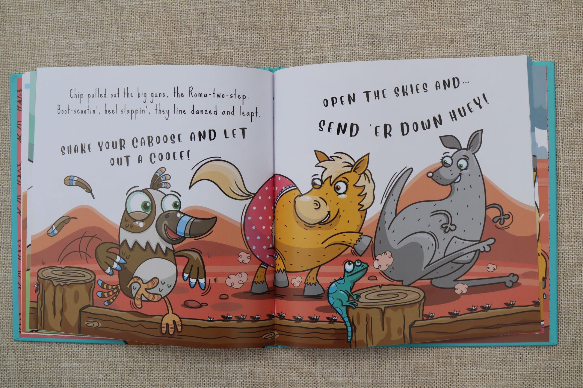 An open Australian children's book spread laid flat on a beige textured fabric background. The illustrated pages show cartoon animals in a desert-like scene with text at the top of each page.