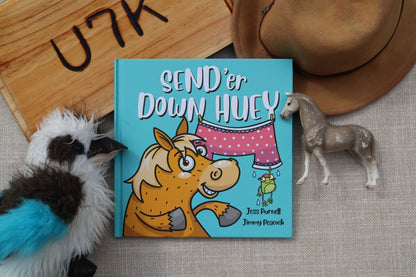 An Australian children's book titled 'SEND 'er Down HUEY' by Australian children's author Jess Purnell and Jimmy Peacock, featuring a cartoon horse and pink polka-dot clothing on a turquoise cover, styled in a flat lay with a toy horse, a plush bird, a hat and a wooden sign.