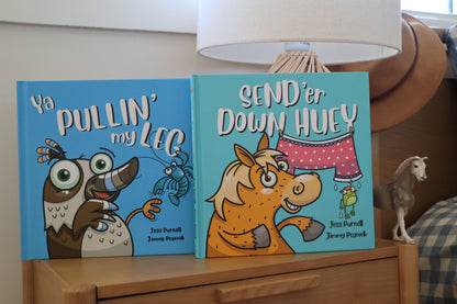 Two Australian children's picture books, 'Ya Pullin' My Leg' and 'Send'er Down Huey', displayed side-by-side on a wooden nightstand next to a lamp.