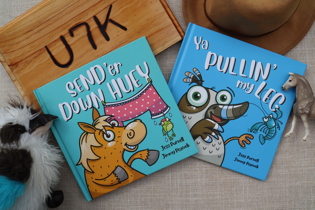Two Australian children's picture books, 'Send'er Down Huey' and 'Ya Pullin' My Leg', displayed on a textured beige fabric alongside a wooden sign, a cowboy hat, and small toy props.