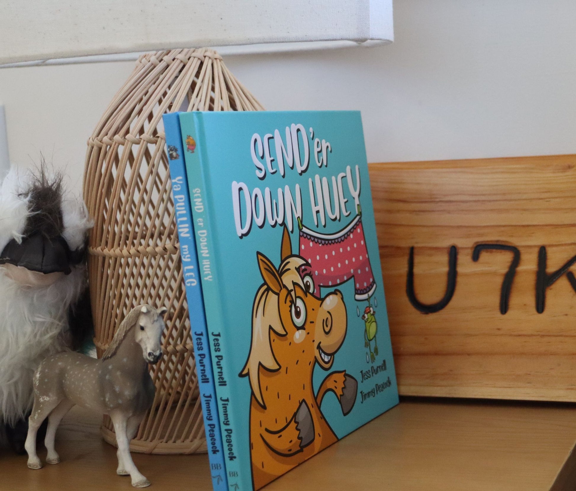 Australian children's book titled "SEND 'er DOWN HUEY" by Jess Purnell and Jimmy Peacock, standing upright on a wooden surface next to a woven basket, a small horse figurine, and a wooden sign.