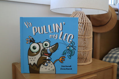 A hardcover Australian children's book titled "Ya PULLIN' my LEG" by Jess Purnell and Jimmy Peacock, standing upright on a wooden bedside table next to a lamp.