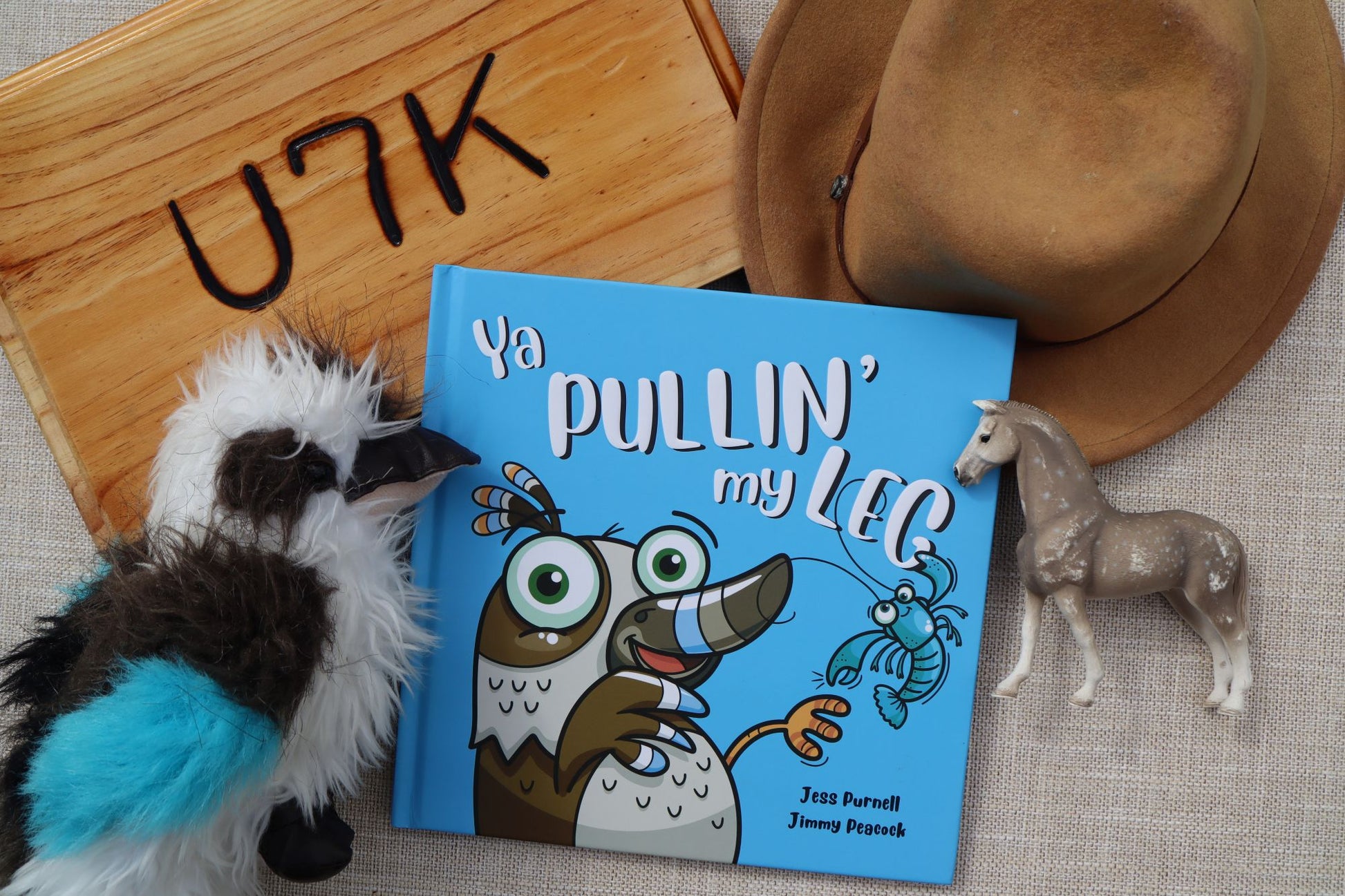 An Australian children's book titled 'Ya PULLIN' my LEG' by Australian children's author Jess Purnell and Jimmy Peacock, styled in a flat-lay with a brown cowboy hat, a small horse figurine, a plush toy, and a wooden sign on a textured fabric background.