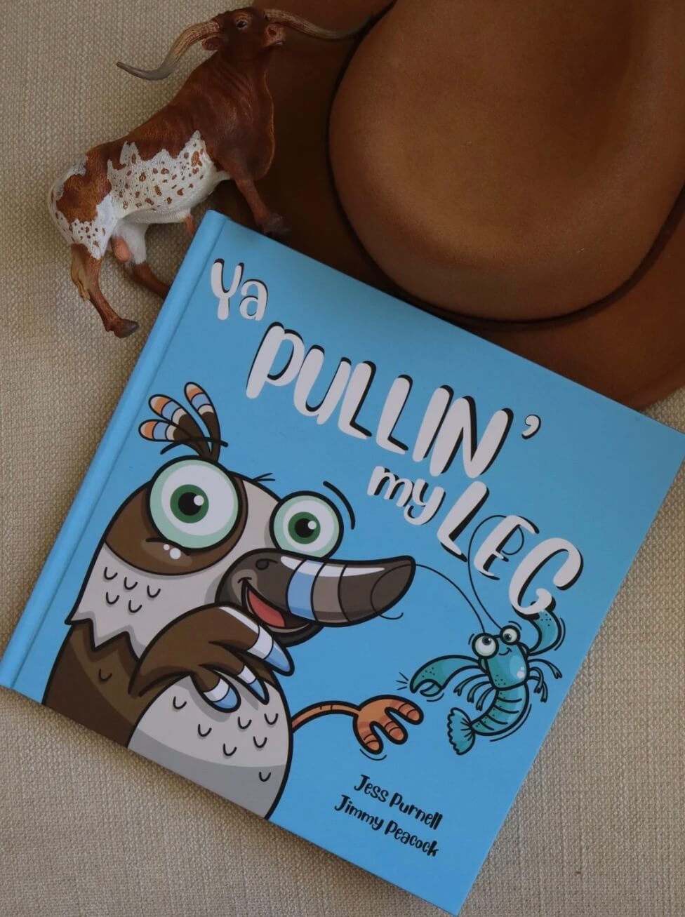 An Australian children's hardcover book titled 'Ya Pullin' My Leg' by Jess Purnell and Jimmy Peacock, featuring a bright blue cover with a cartoon bird and crab. The book is styled on a beige textured surface next to a brown cowboy hat and a small bull figurine.