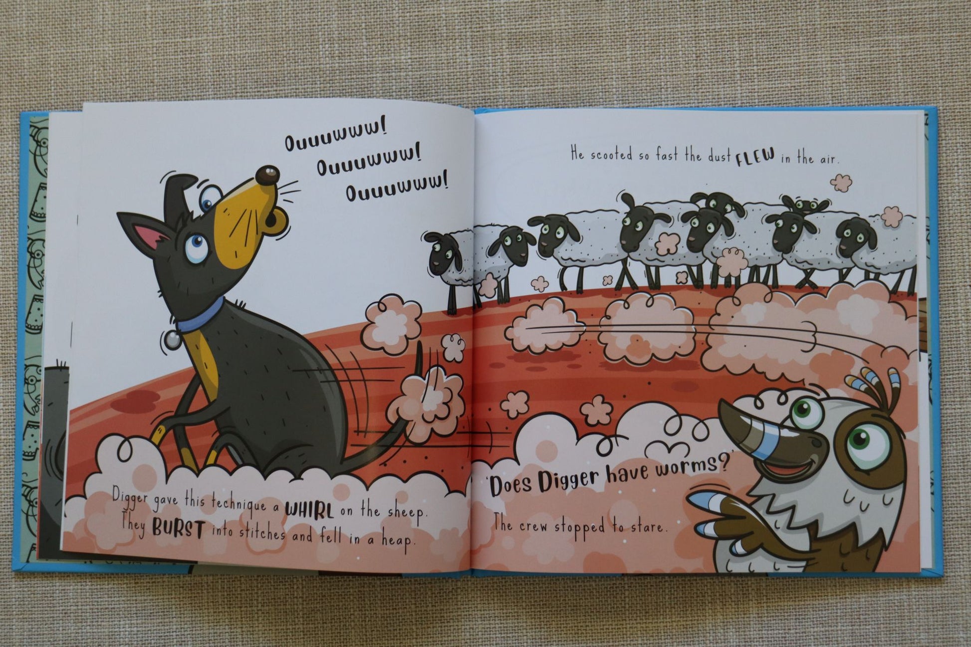 An open children's book spread laid on a beige textured fabric background. The illustrated pages show a cartoon dog and a bird with text and a worm-themed scene.