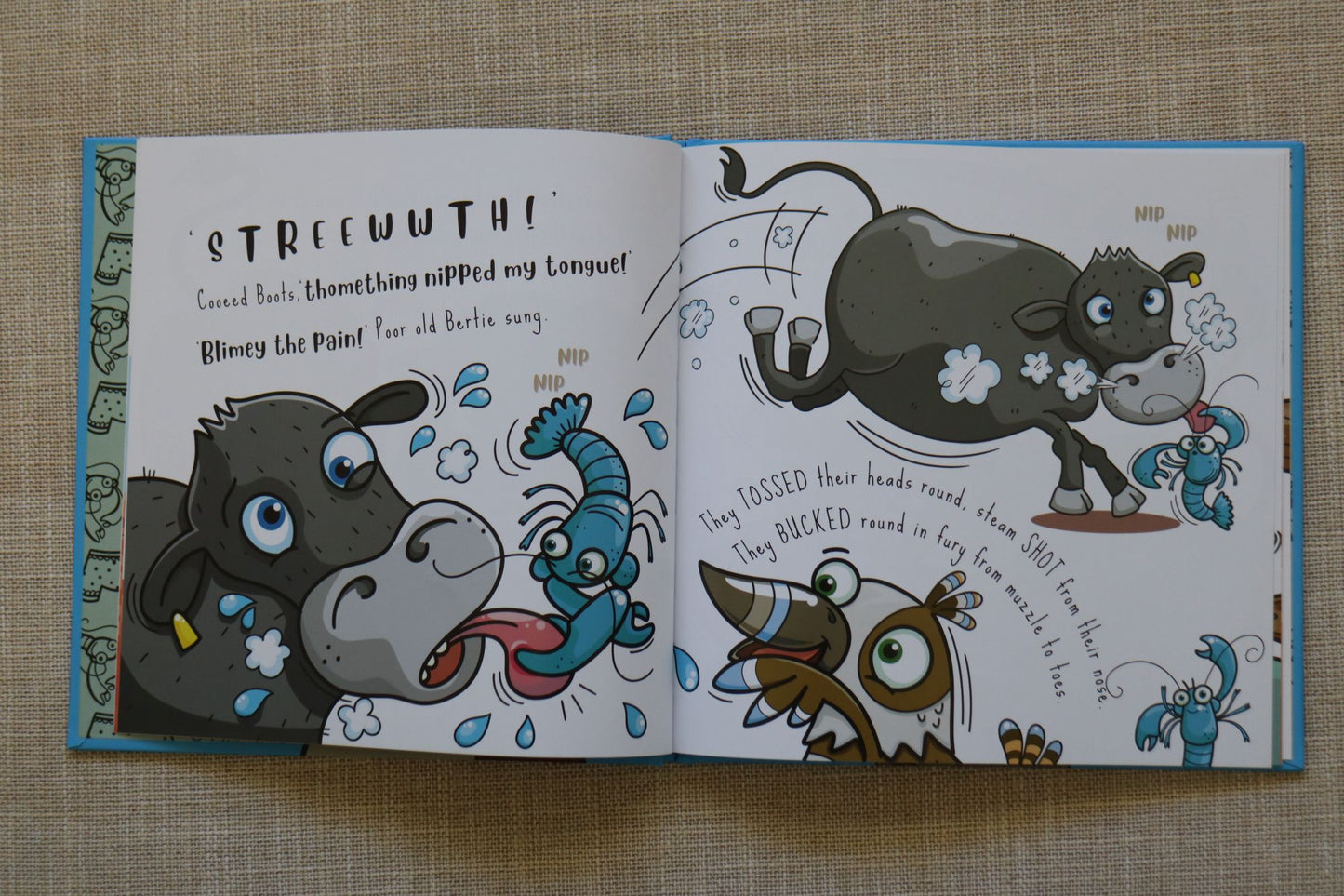 An open Australian children's book spread laid flat on a beige textured fabric background, featuring colorful cartoon animal illustrations and printed text. The pages show a dark gray steer, blue yabbies and a kookaburra in a playful scene.