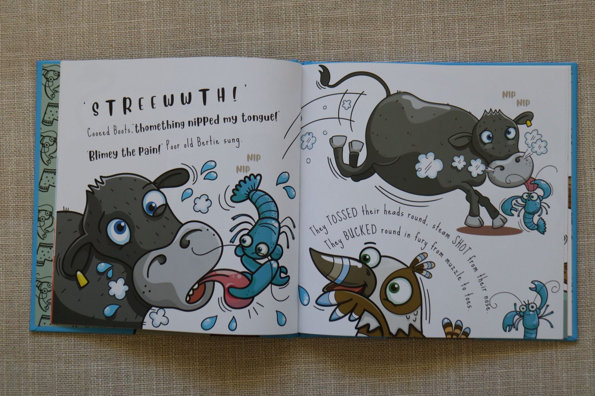 An open Australian children's book spread laid flat on a beige textured fabric background, featuring colorful cartoon animal illustrations and printed text. The pages show a dark gray steer, blue yabbies and a kookaburra in a playful scene.